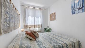 Een slaapkamer met een bed, een raam met dunne gordijnen, een kunstwerk aan de muur en een kamerplant.