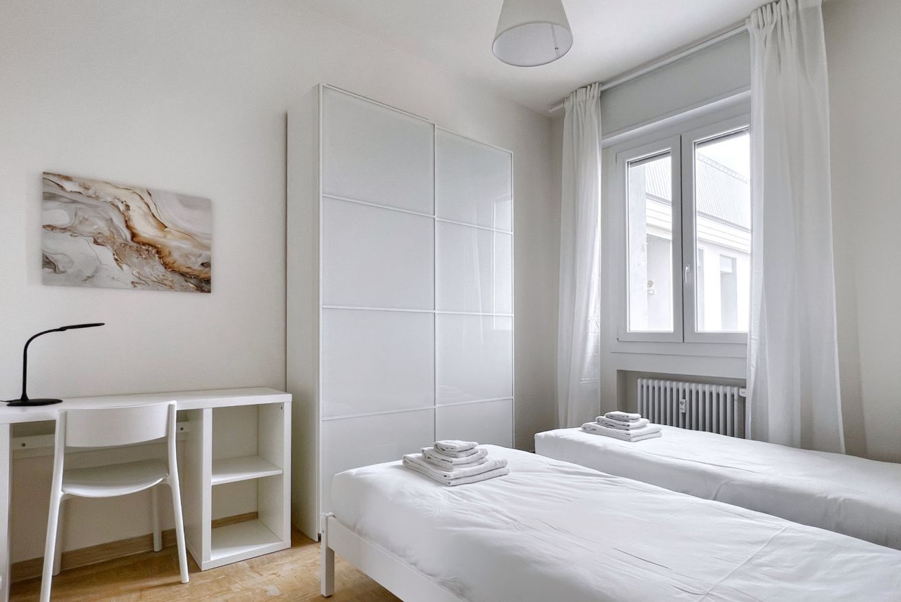 A hotel room with a bed, a desk, a chair, a radiator, and a window with curtains.