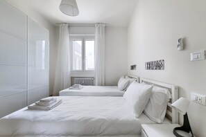 Habitació d'hotel amb dos llits, una finestra amb cortines, un radiador, una làmpada penjada a la paret i una presa elèctrica.