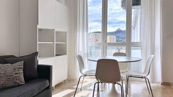 A modern living room with a sofa, a dining table with chairs, a white built-in shelving unit, and a large window with a view of buildings and a hill.