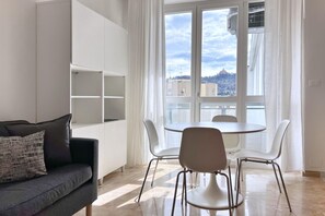 Ein modernes Wohnzimmer mit einer Couch, einem Esstisch mit Stühlen, einer weißen, eingelassenen Regaleinrichtung und einem großen Fenster mit Blick auf Gebäude und eine Hügelkulisse.