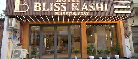 A hotel named 'BLISS KASHI' with a modern facade, large glass entrance, and potted plants at the entrance.