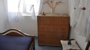 A bedroom with a wooden bed, a dresser with a vase of dried plants, and a window with sheer curtains.