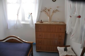 A bedroom with a wooden bed, a dresser with a vase of dried plants, and a window with sheer curtains.