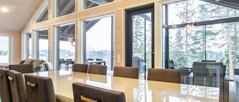 Un vaste espace dédié à la salle à manger, avec une longue table et de nombreuses chaises, de grandes fenêtres offrant une vue sur l’extérieur et un lustre suspendu au plafond.