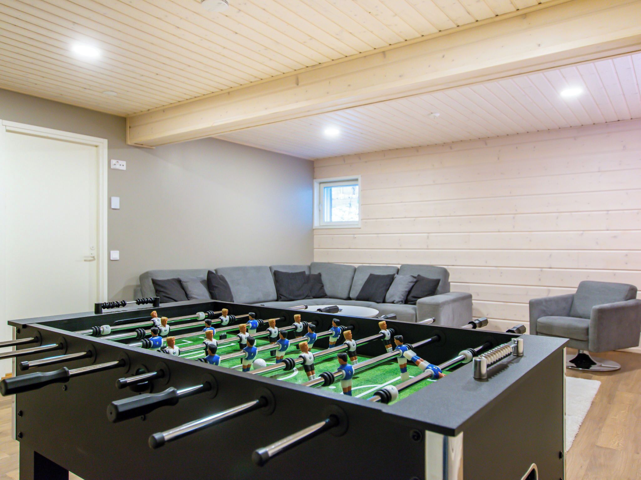 A room with a foosball table, a grey sofa, a grey armchair, and a window with daylight.