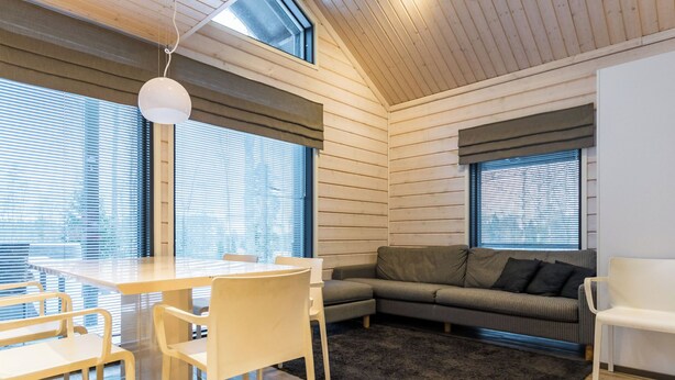 A modern interior with a wooden ceiling, a dining area with a table and chairs, a sofa, and large windows with blinds.