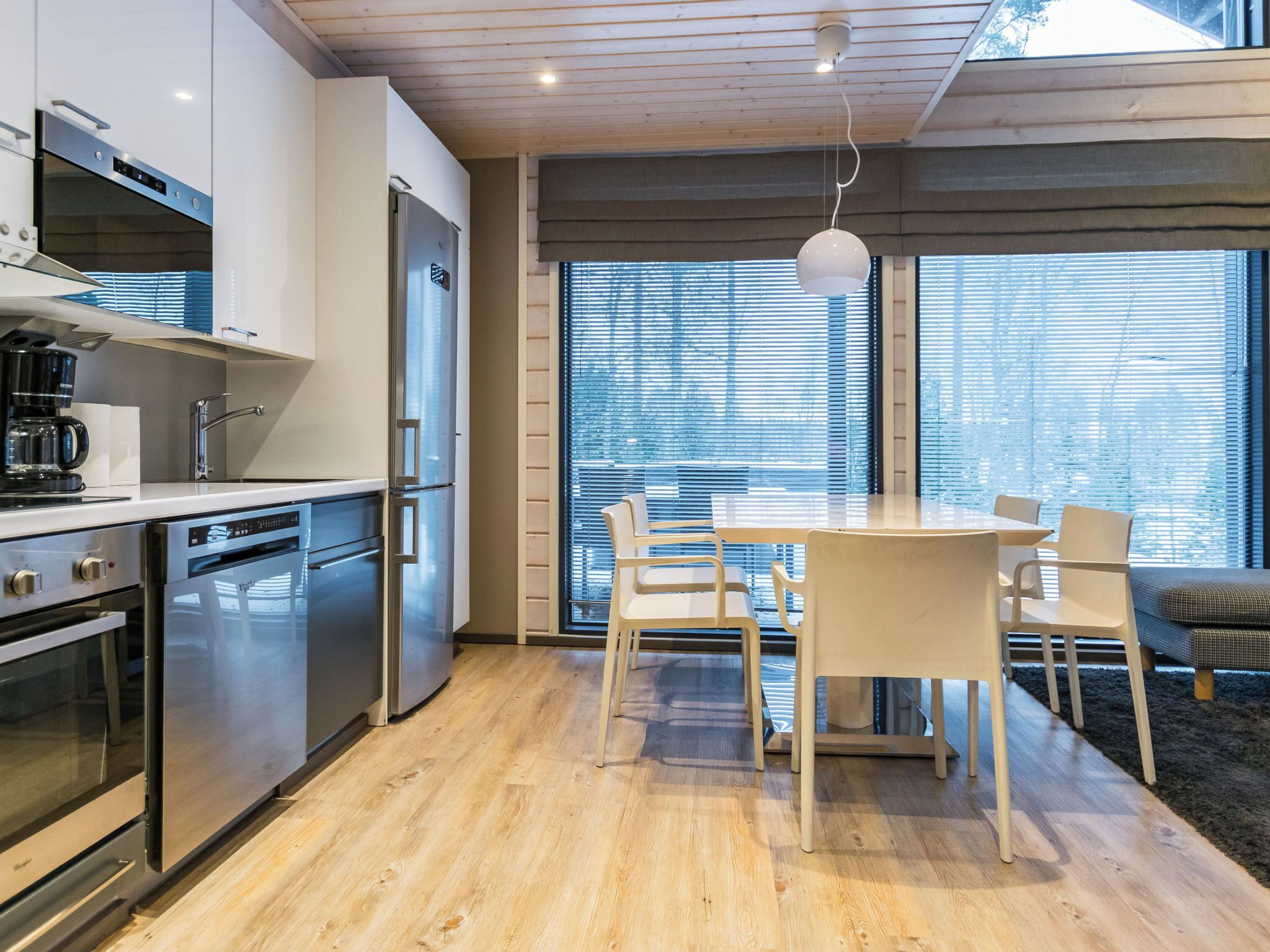 A modern kitchen with stainless steel appliances, a dining area with a table and chairs, and large windows with blinds.
