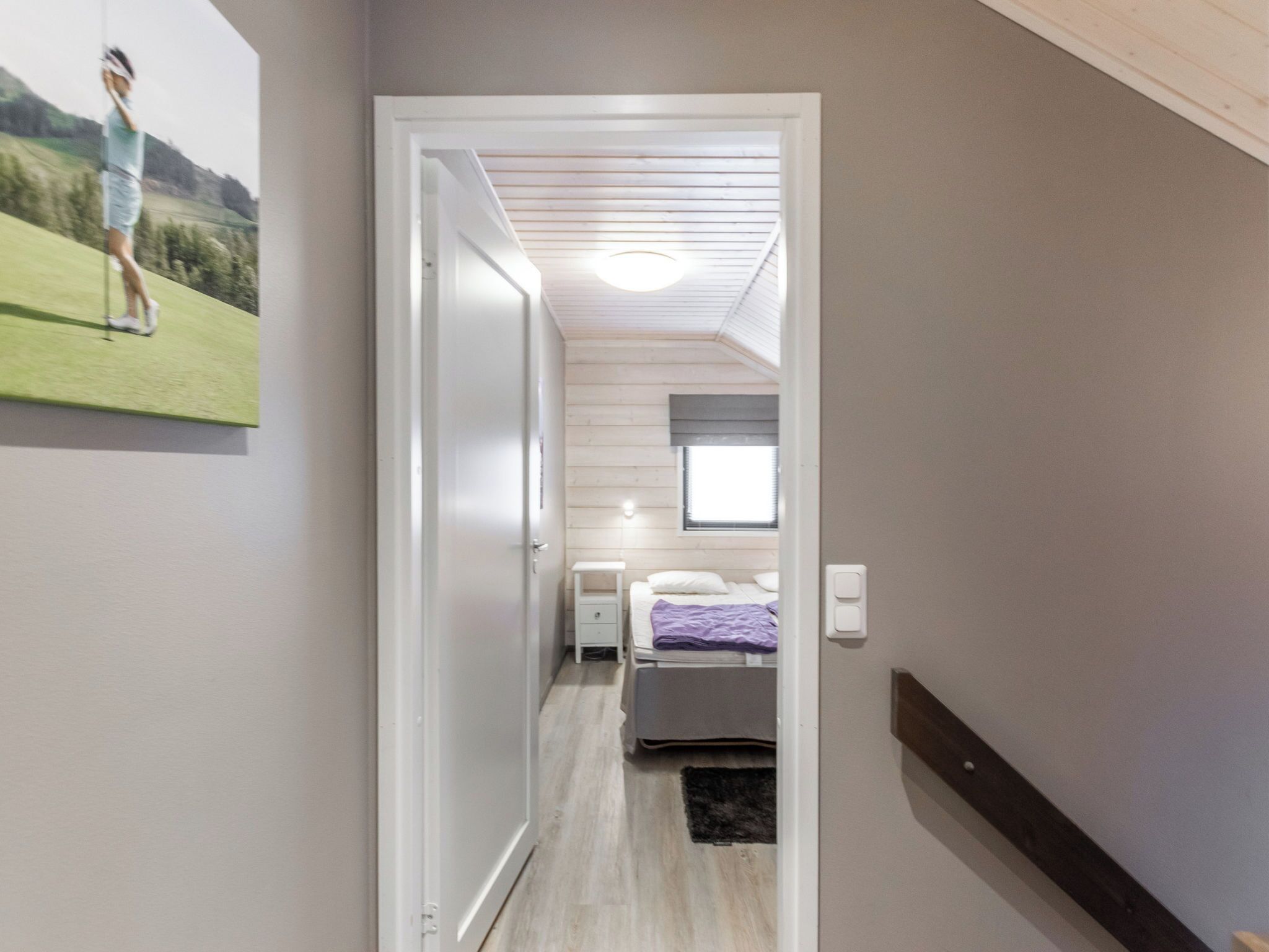 Een smalle gang die uitkomt in een slaapkamer, met een bed, nachtkastje en een raam.