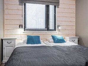 A bedroom with a bed, two bedside tables, a window with blinds, and a wall with horizontal wooden panels.