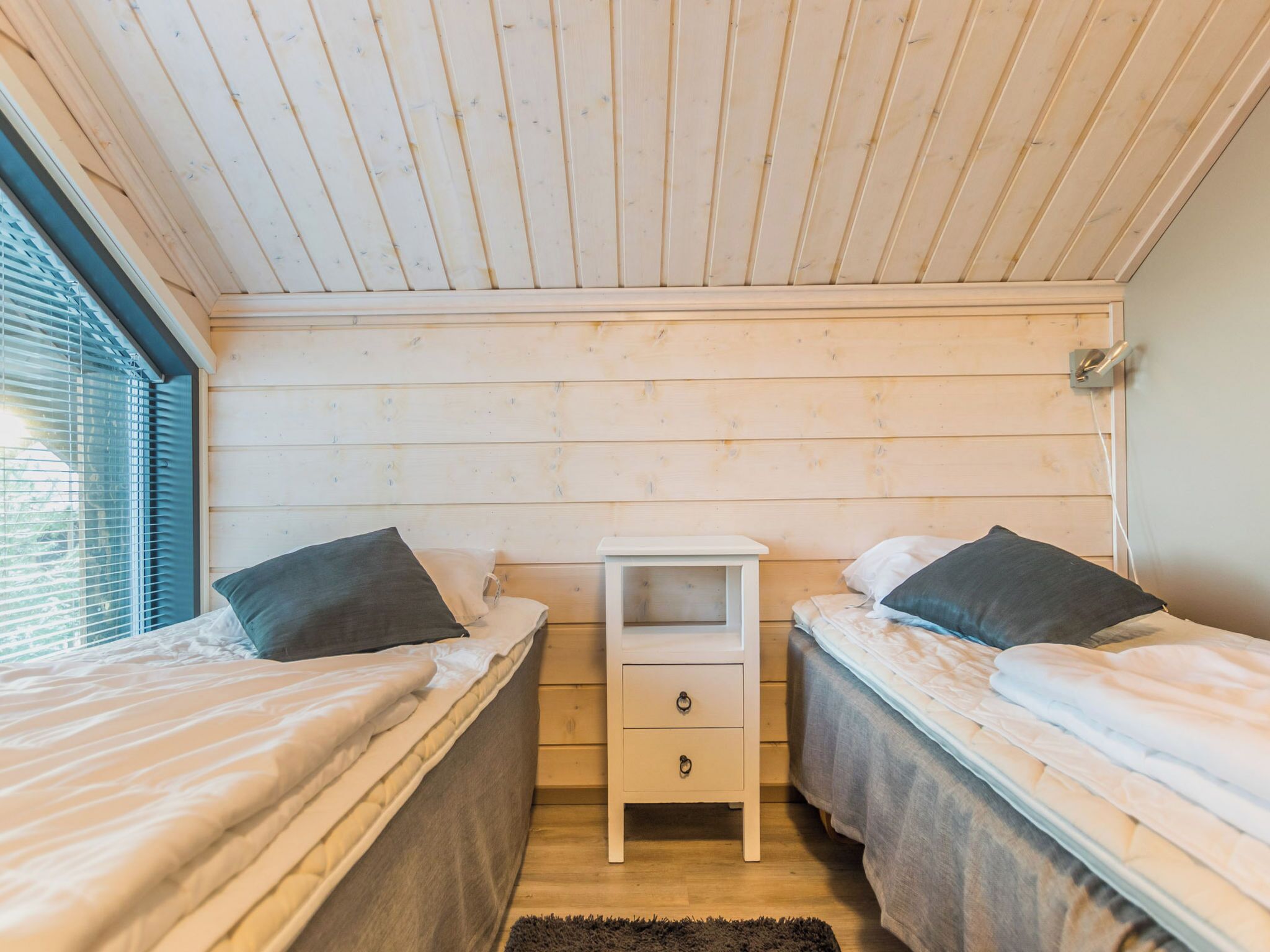 Een kamer met twee bedden, een klein nachtkastje met lades, een raam met zonwering, en een houten plafond.