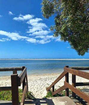 Un banco de madera que conduce a una playa de arena, bajo un cielo azul despejado y un mar tranquilo.