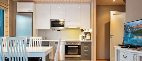 Une cuisine moderne avec des armoires blanches, un four encastré et un four à micro-ondes. On y trouve une table à manger avec des chaises, un téléviseur posé sur un meuble et un plancher en bois.