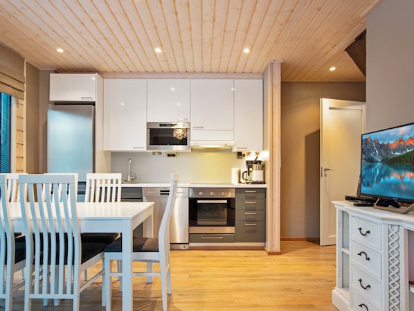 A modern kitchen with white cabinets, a built-in oven, and a microwave. There is a dining table with chairs, a TV on a stand, and a wooden floor.