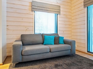 A grey sofa with two teal cushions in a room with wooden walls and a window with blinds.