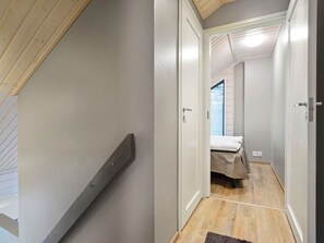 A narrow hallway with a wooden staircase on the left, a bed with a white mattress and a beige blanket in the center, and a door on the right.