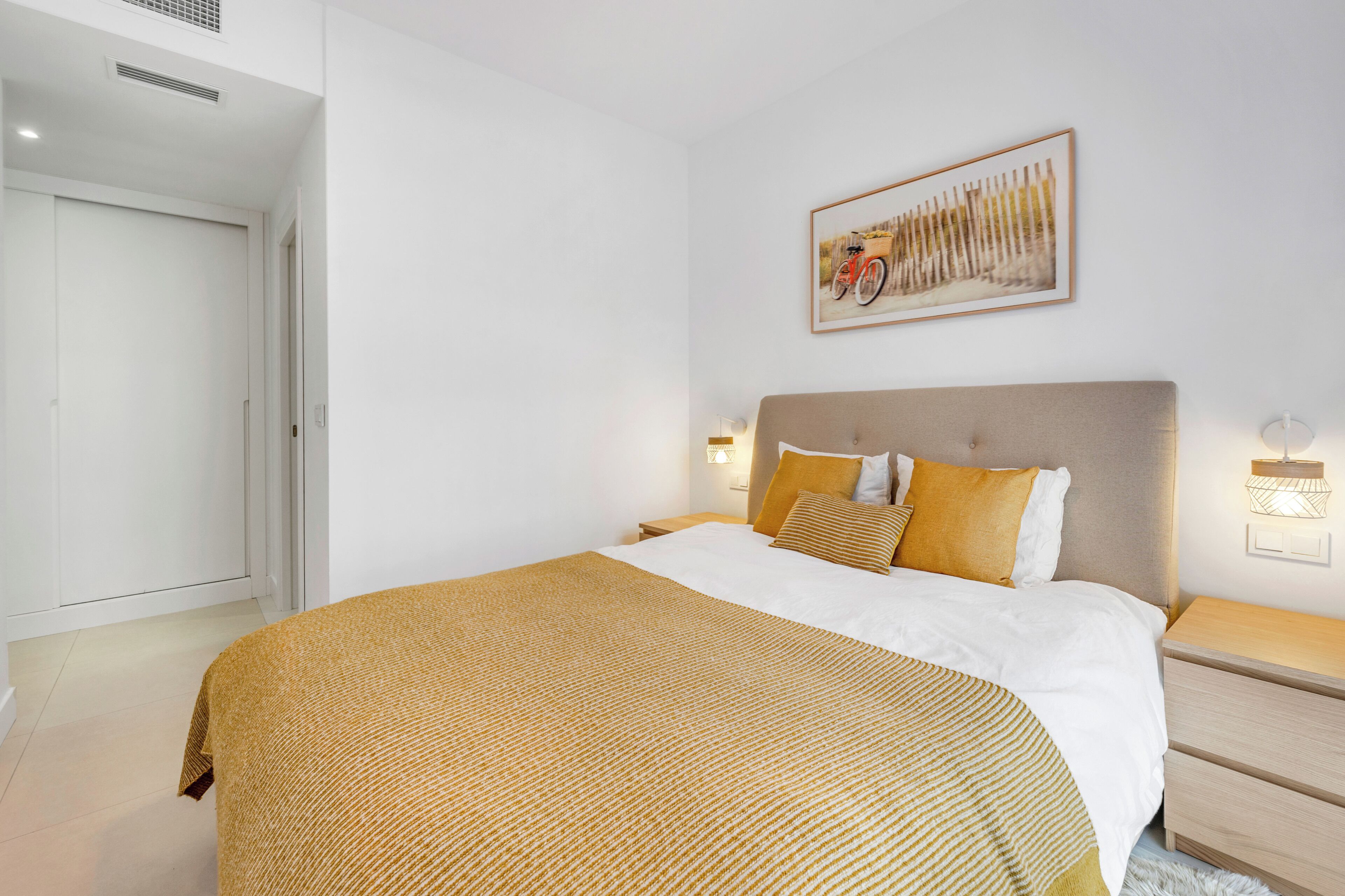 A bedroom with a bed, a nightstand, a wall-mounted light, a door, and a framed picture on the wall.