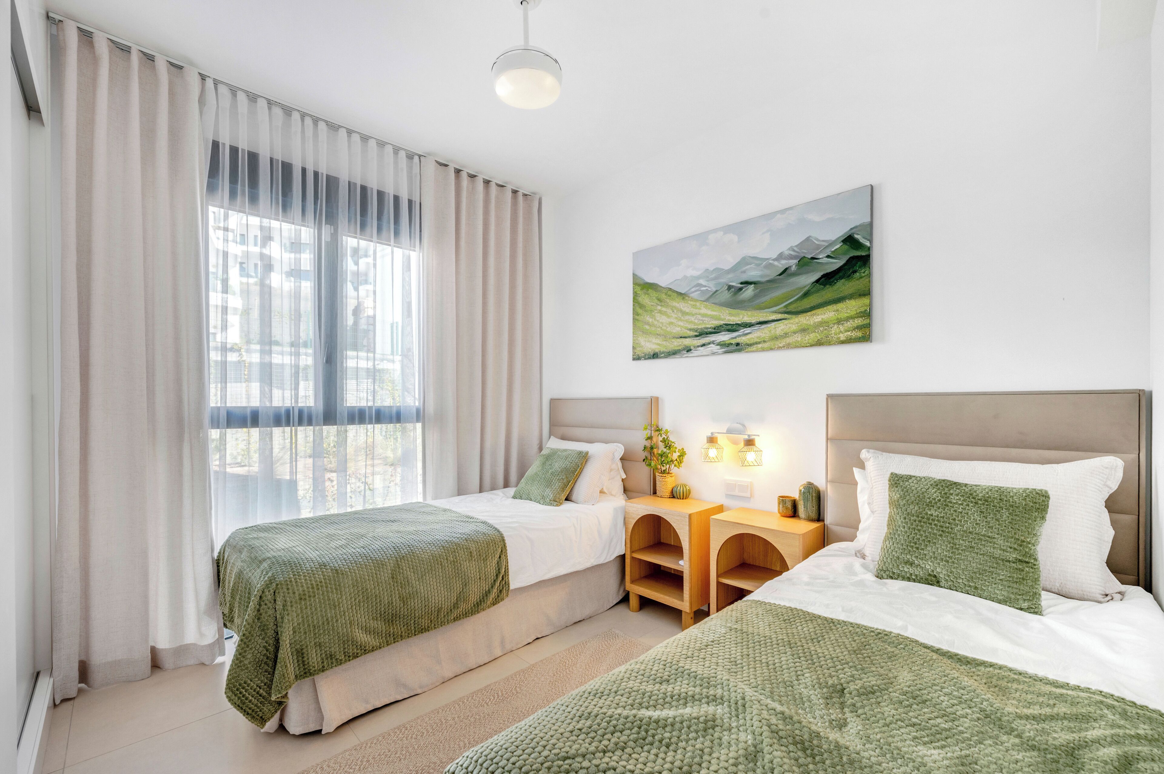A hotel room with two beds, a large window with sheer curtains, a painting of a landscape, and a bedside table with a lamp.