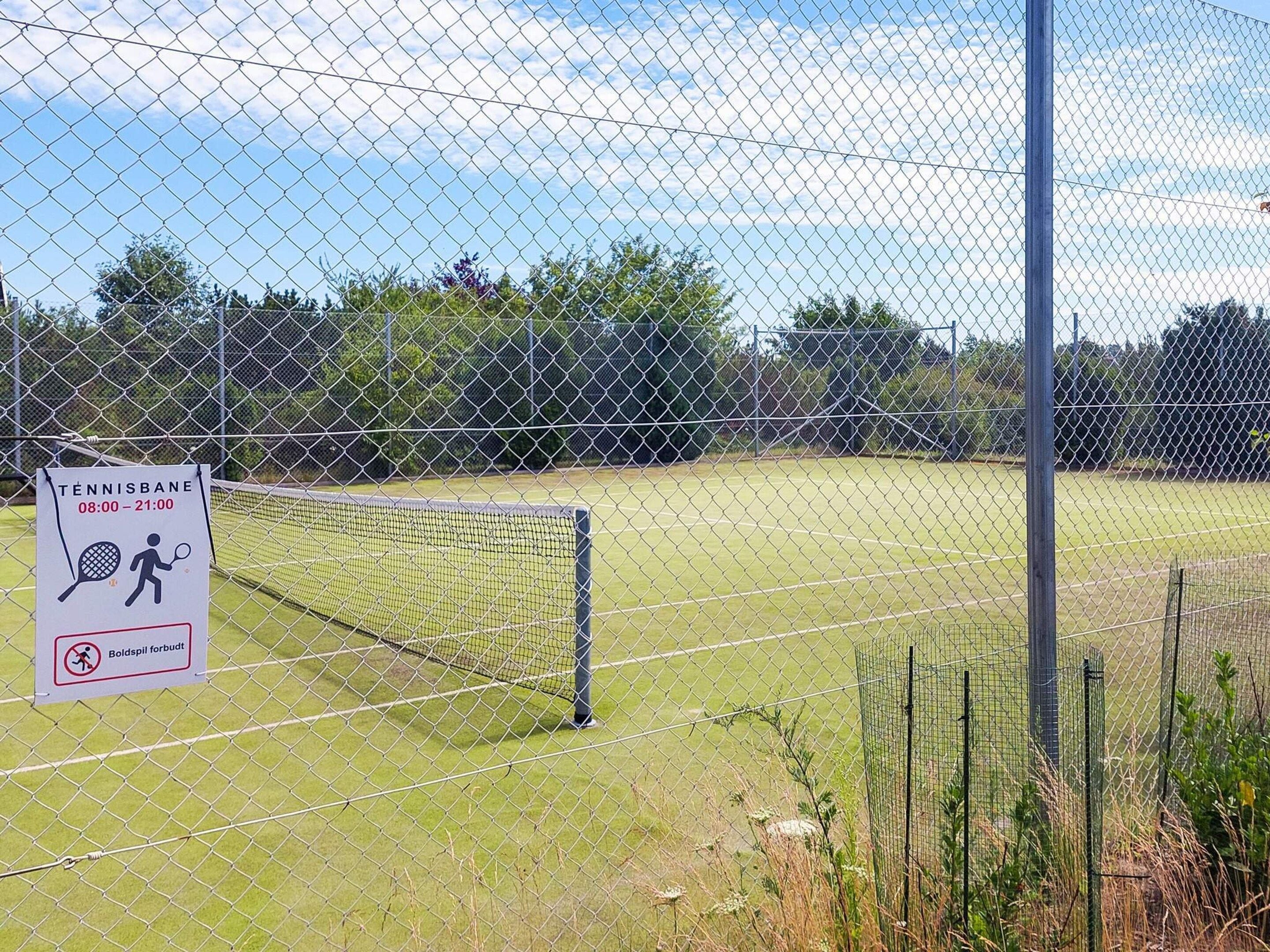 A tennis court with a chain-link fence, a sign indicating the court's name and operating hours, and a no ballspotting warning.