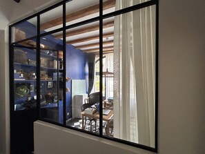 A room with a large window, a bookshelf, a desk with a computer, and a chair.