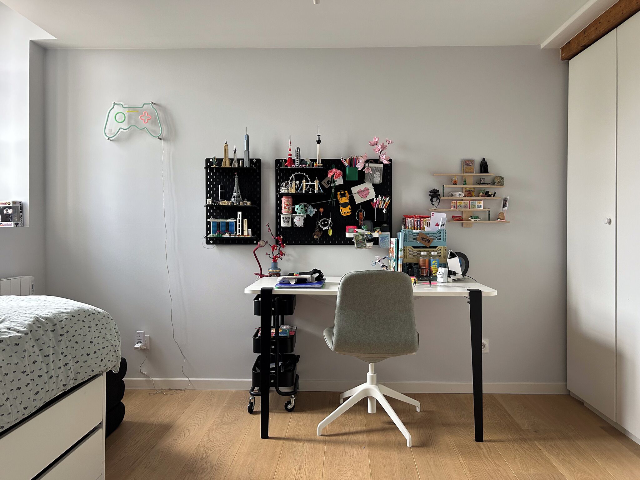 A bedroom with a bed, a desk with a chair, a wall-mounted shelf with various items, and a neon game controller on the wall.