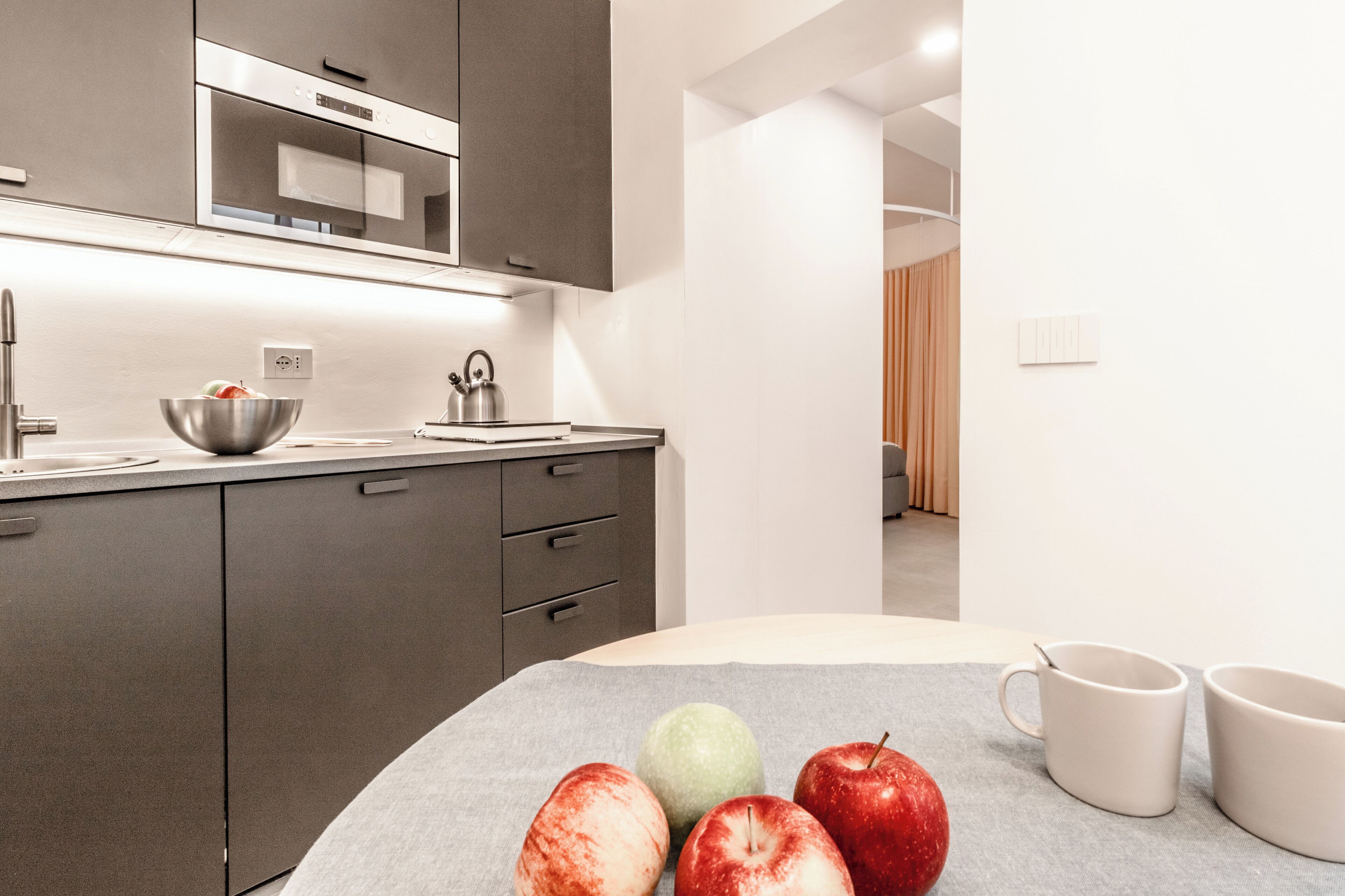 Une cuisine moderne équipée d’un four à micro-ondes, d’une bouilloire et d’un saladier rempli de fruits, posé sur le plan de travail.