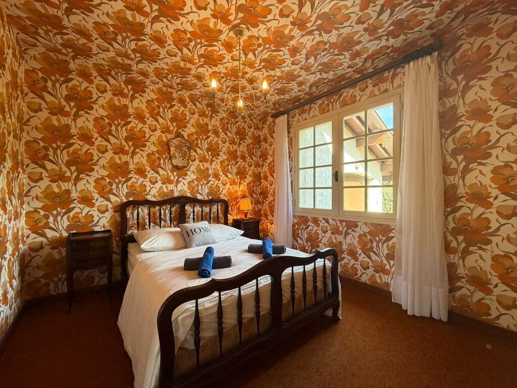 Ein Schlafzimmer mit floralem Wandpapier, einem hölzernen Bett, einem Nachttisch und einem Fenster mit Vorhängen.