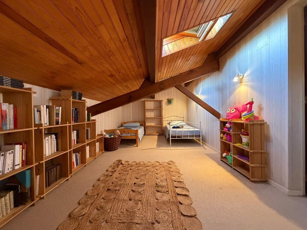 Ein Zimmer mit schrägen Wänden, hölzernen Bücherregalen, einem gewebten Teppich und einem Bett mit blauer Matratze.