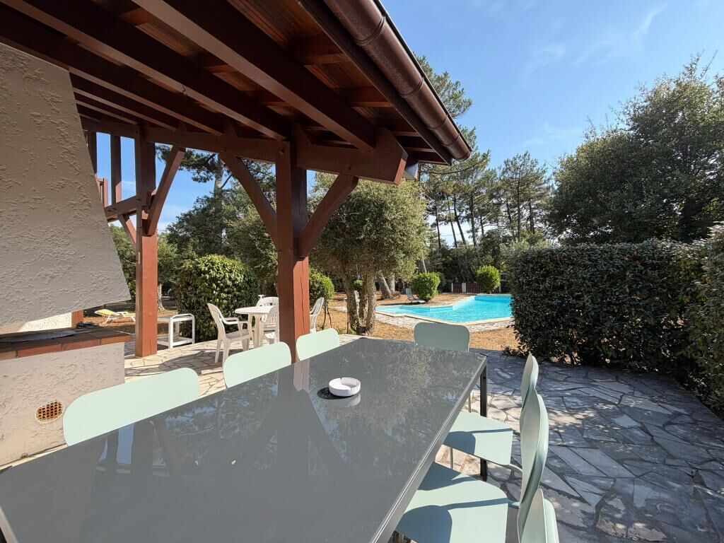 Un espace extérieur aménagé en terrasse, avec une table et des chaises, une piscine en arrière-plan et une structure en pergola en bois.