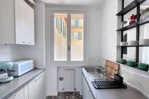 Ett litet kök med ett fönster, en mikrovågsugn, en diskho och en hylla med koppar.