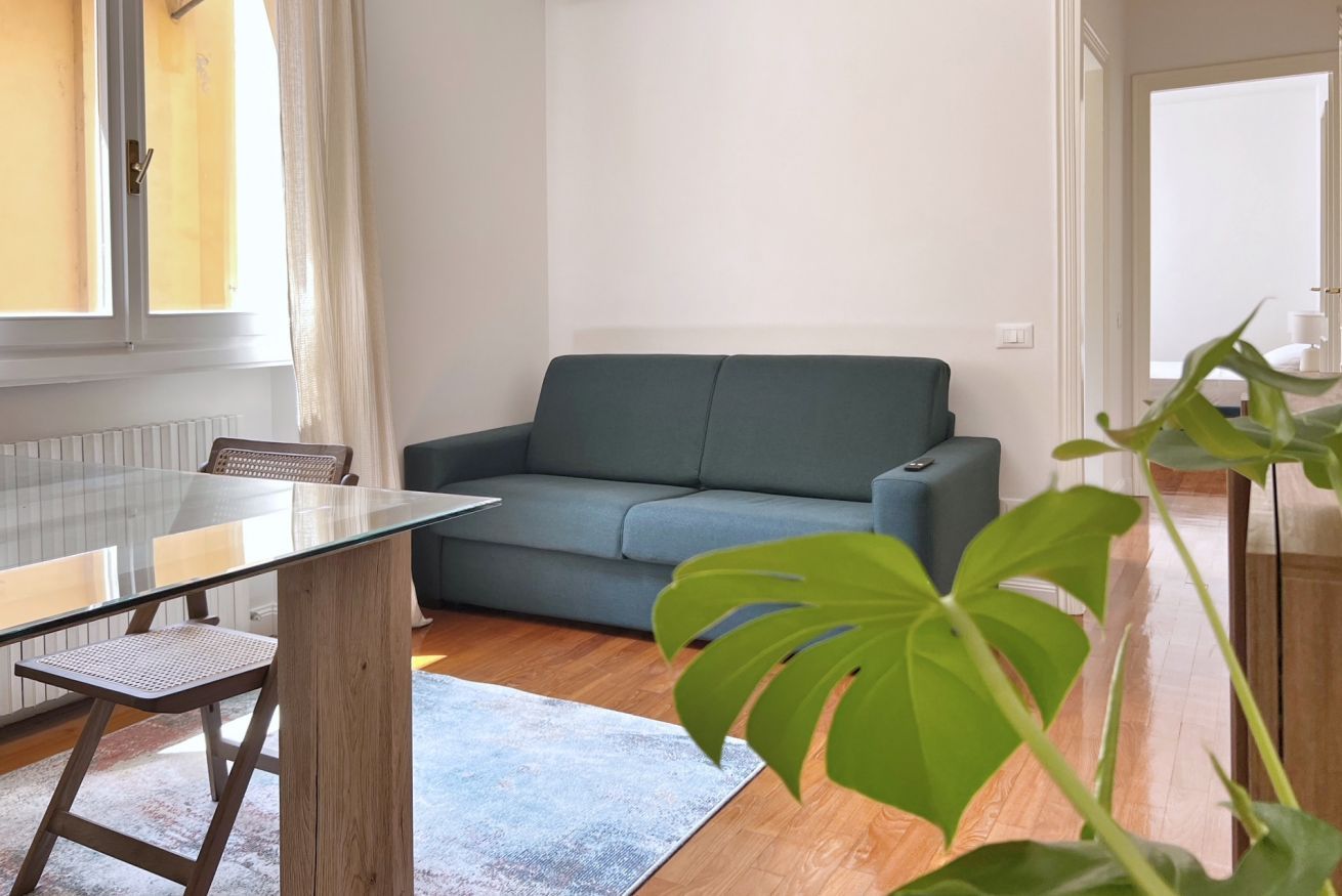 A living room with a glass-top table, a grey sofa, a wooden floor, and a large green plant.