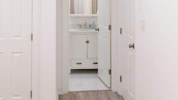 Un couloir étroit mène à une salle de bain équipée d’un miroir, d’un lavabo et de placards.