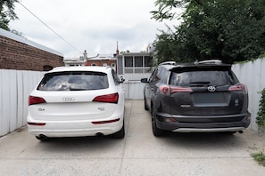 Two parked cars, a white sedan and a black SUV, in a narrow driveway with a white fence on one side and a brick wall on the other. - A stylish retreat in Mount Pleasant, one of DC's most walkable neighborhoods. Two bedrooms plus a sofa bed sleep six, with two full baths - plus the city's rarest luxury: your own private off-street parking. Walk to Metro, cafes & Zoo. (Washington)