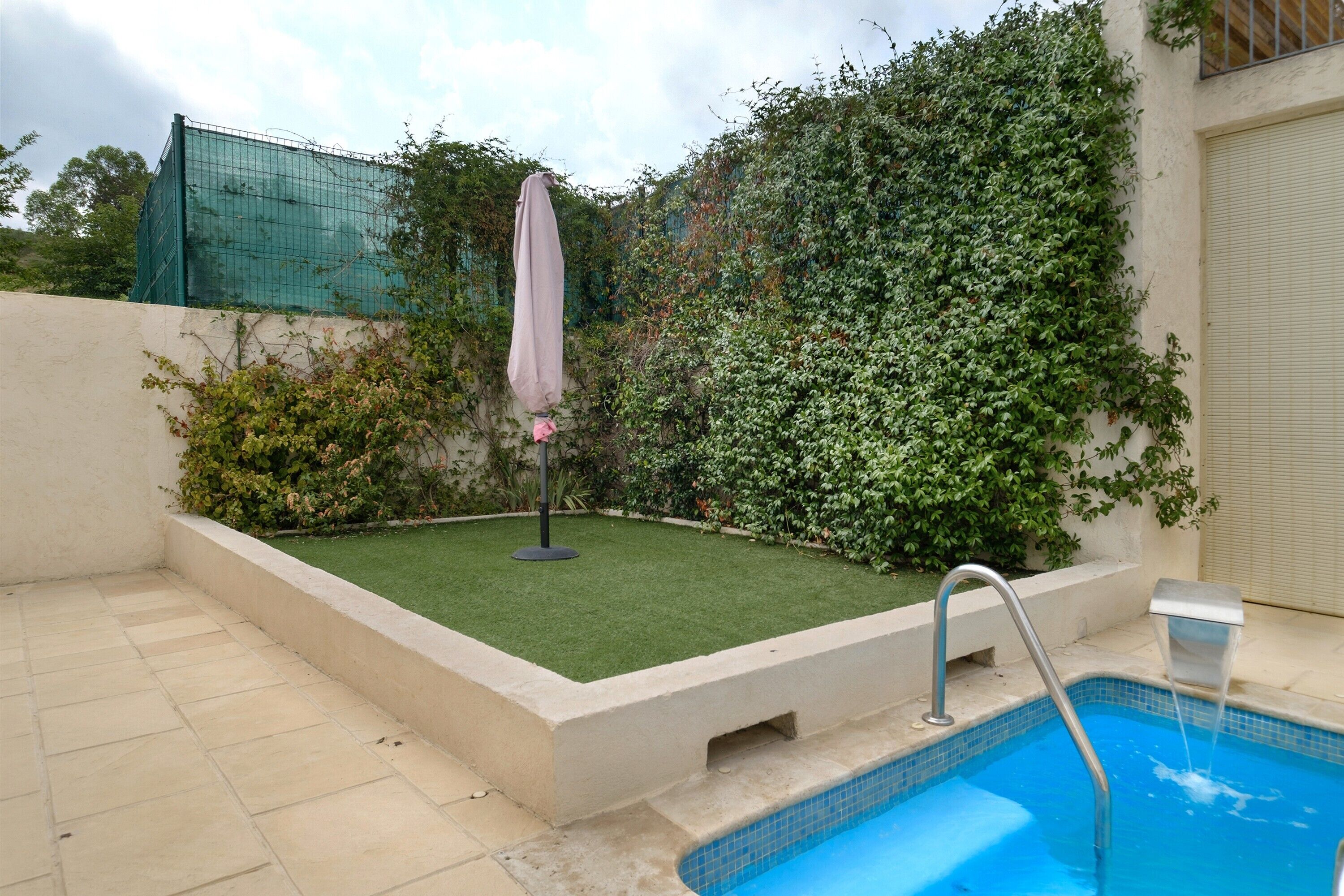 Une piscine avec une terrasse carrelée, un petit jardin agrémenté de gazon synthétique et une clôture en métal.