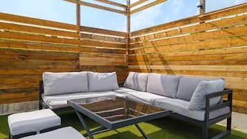A covered outdoor seating area with a sectional sofa and a glass table, surrounded by wooden slats and artificial grass.