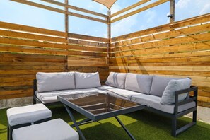 Ein überdachter Außenbereich mit Ecksofa und Glastisch, umgeben von Holzlatten und künstlichem Rasen.