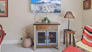 Ein Wohnzimmer mit an der Wand montiertem Fernseher, einer Kommode mit Schubladen, einem Korbsessel, einer Lampe und einer Vase mit Trockenpflanzen.