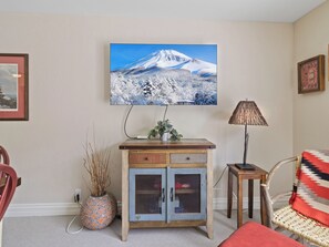 Ein Wohnzimmer mit an der Wand montiertem Fernseher, einer Kommode mit Schubladen, einem Korbsessel, einer Lampe und einer Vase mit Trockenpflanzen.