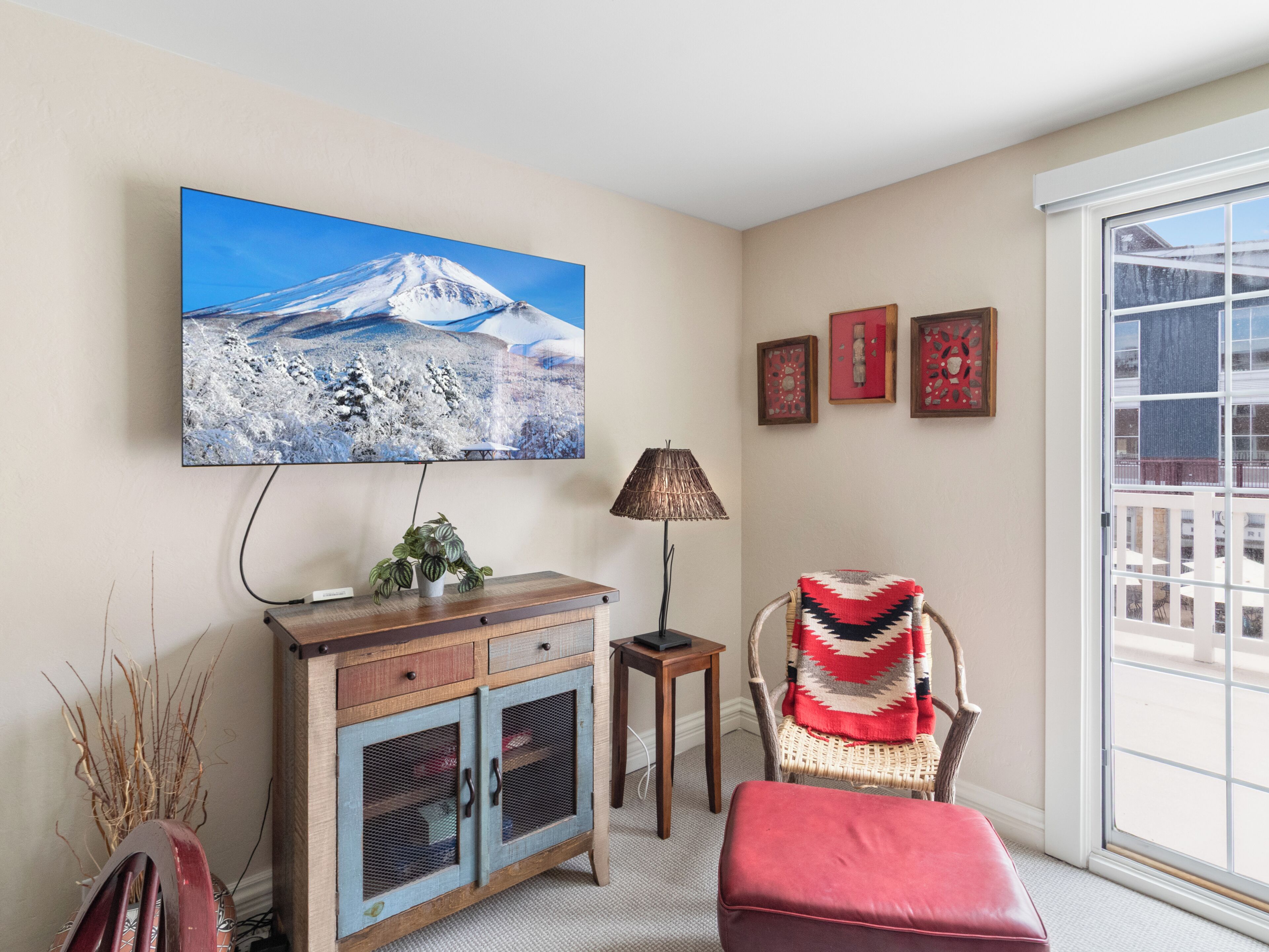 A living room with a television mounted on the wall, a cabinet, a chair with a blanket, and a window with a view.