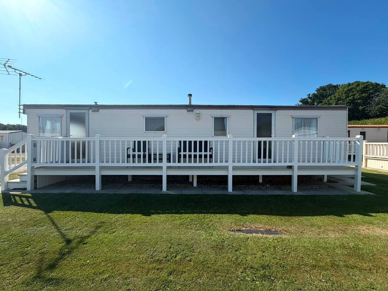 Ein eingeschossiges Mobilheim mit weißer Fassade, einer Veranda mit weißen Geländern und einer kleinen angebauten Garage.