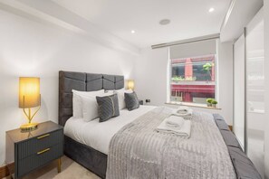 A modern bedroom with a large bed, bedside lamps, a nightstand, and a window with a view of buildings. - Marylebone's Gold - 3 Bed (Greater London)