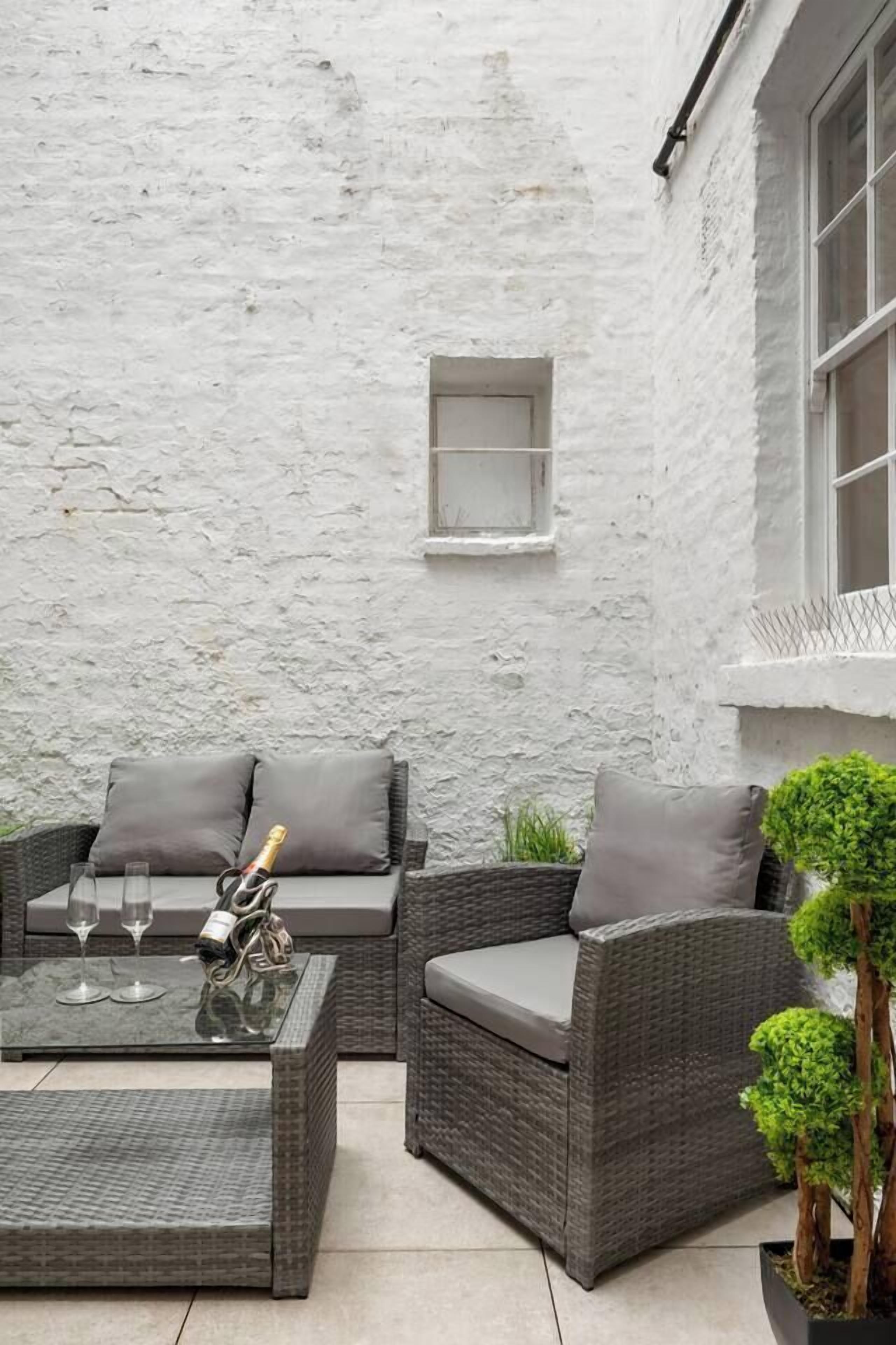 An outdoor seating area with wicker furniture and a glass table, a white textured wall, and a potted plant.