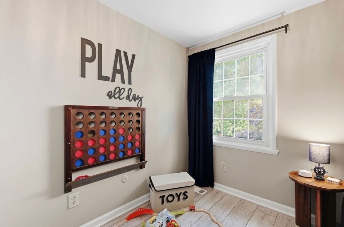 A room with a wall-mounted game board, a window with curtains, a small table with a lamp, and a box labeled 'TOYS'.