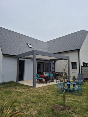 Et moderne hus med en overdækket terrasse, udendørs møbler og et lille træ.