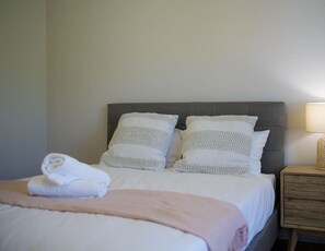 Ein ordentlich bezogenes Bett mit Kissen und einem gefalteten Handtuch, eine Nachttischlampe sowie ein Nachttisch aus Holz.