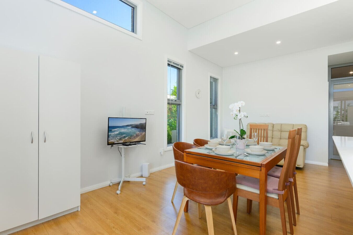 Uma sala de jantar moderna com uma mesa de madeira posta para quatro pessoas, uma televisão em um suporte e vista para o exterior pela janela.