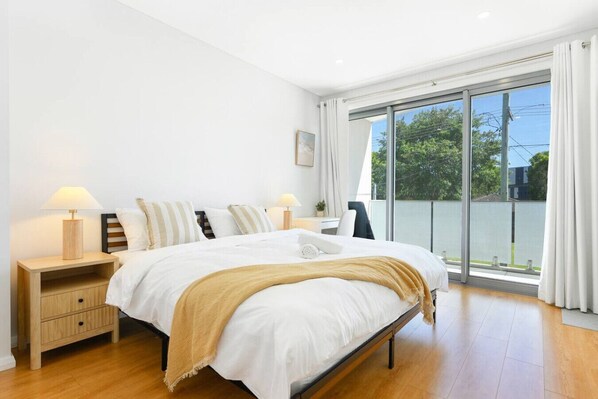 A modern bedroom with a large bed, two bedside tables, a chair, and a sliding glass door leading to a balcony.