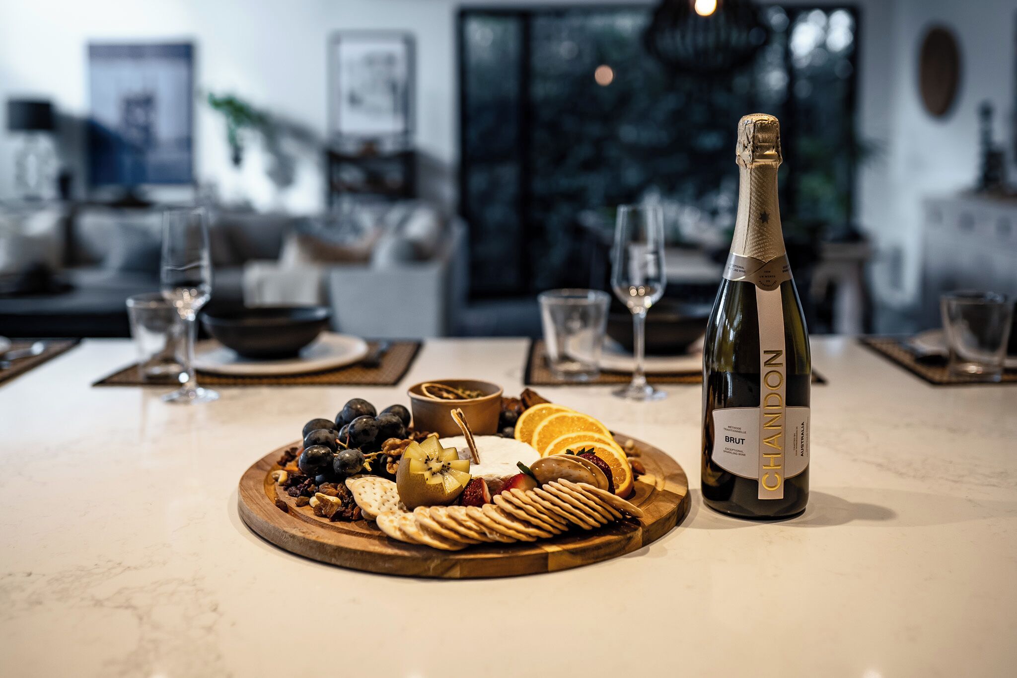 Ein Holzbrett mit einer Auswahl an Käse, Früchten und Crackern, begleitet von einer Champagnerflasche.