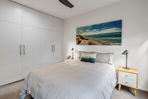 Ein Schlafzimmer mit einem Bett, einem Nachttisch, einer Lampe, einem Kleiderschrank und einem Wandbild mit Strandmotiv.
