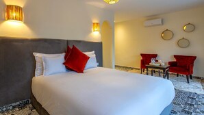 A hotel room with a large bed, two red chairs, a small table, and a decorative wall hanging.